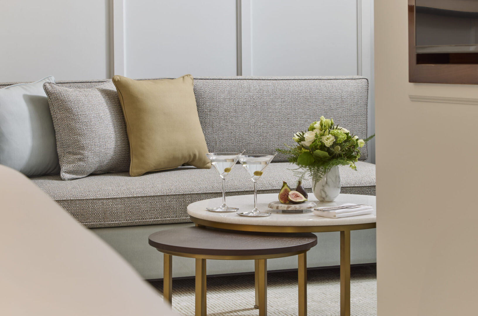 Elegant hotel suite sitting area at The Hawthorn by Galway Bay, with a grey upholstered sofa, gold accent cushions, martini glasses, fresh florals, and nesting gold and marble coffee tables