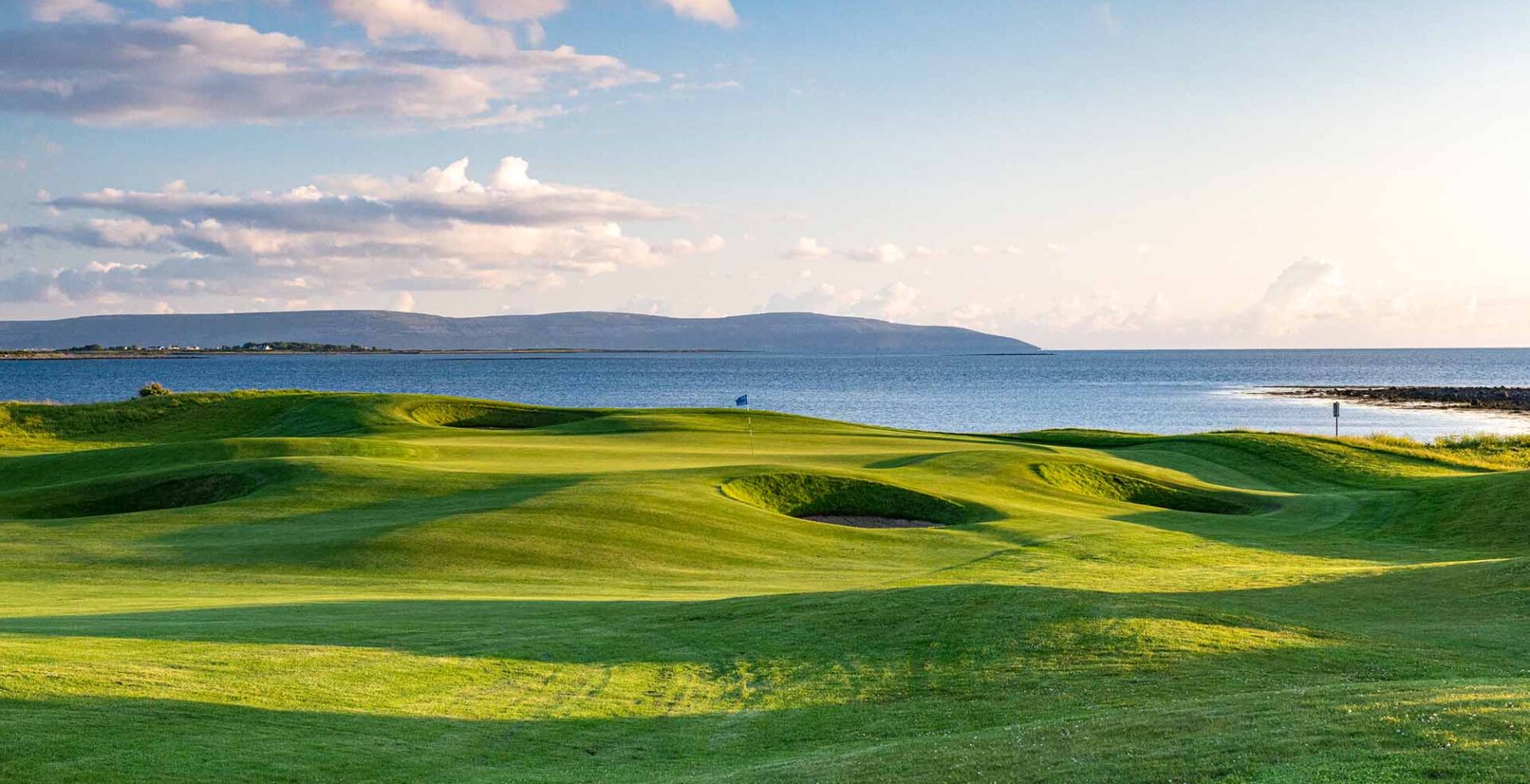 Coastal links golf course at The Hawthorn by Galway Bay, with undulating emerald fairways, a calm blue bay, and the hills of County Clare visible across the water in golden evening light