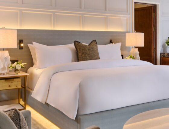 Luxury hotel king bedroom at The Hawthorn by Galway Bay, featuring crisp white linens, grey upholstered headboard with warm cove lighting, textured accent pillow, sculptural bedside lamps, gold brass nightstands, and fresh white rose arrangements